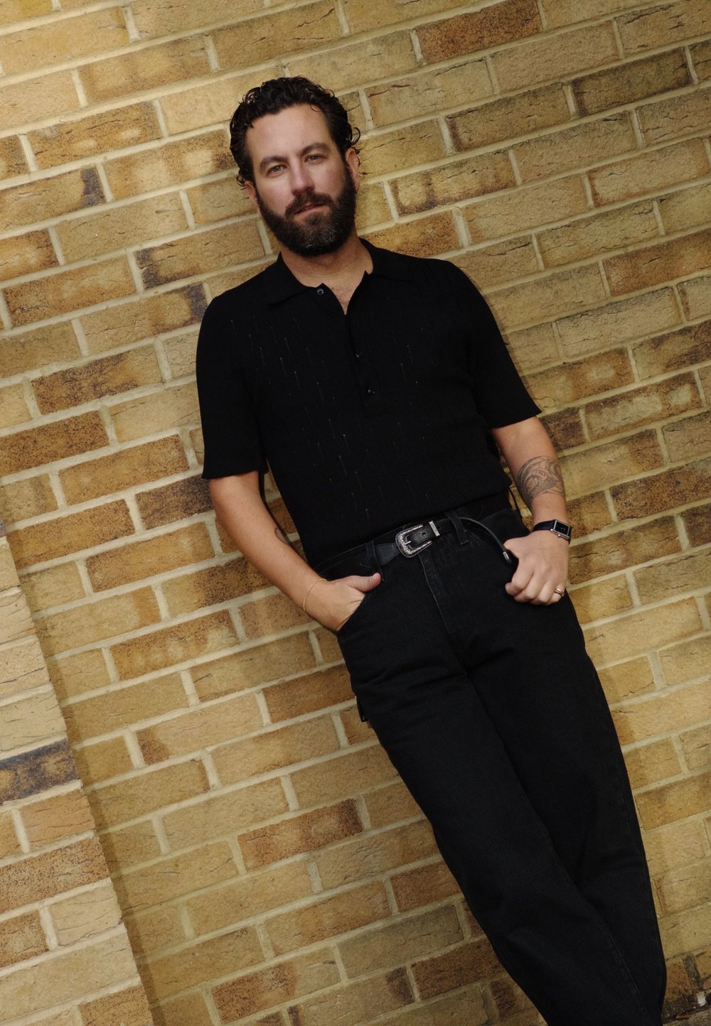 Μαύρο πόλο μπλουζάκι με υφή ζακάρ, κοντά μανίκια και κουμπιά, φοριέται με ψηλόμεσα μαύρα τζιν και ασημένια ζώνη. Φόντο με τοίχο από τούβλα.