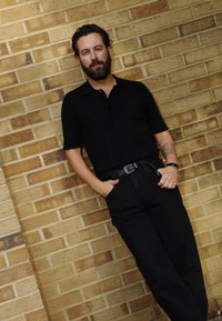 Μαύρο πόλο μπλουζάκι με υφή ζακάρ, κοντά μανίκια και κουμπιά, φοριέται με ψηλόμεσα μαύρα τζιν και ασημένια ζώνη. Φόντο με τοίχο από τούβλα.