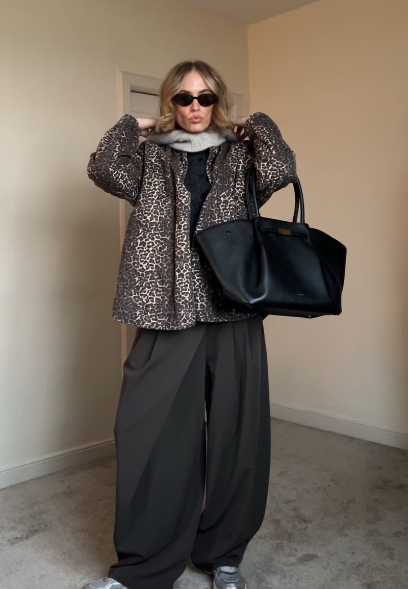 Femme ajustant son col, portant une veste à imprimé léopard, un pantalon large noir, des lunettes de soleil noires, une écharpe grise, des chaussures argentées, tenant un grand sac à main noir à l'intérieur.