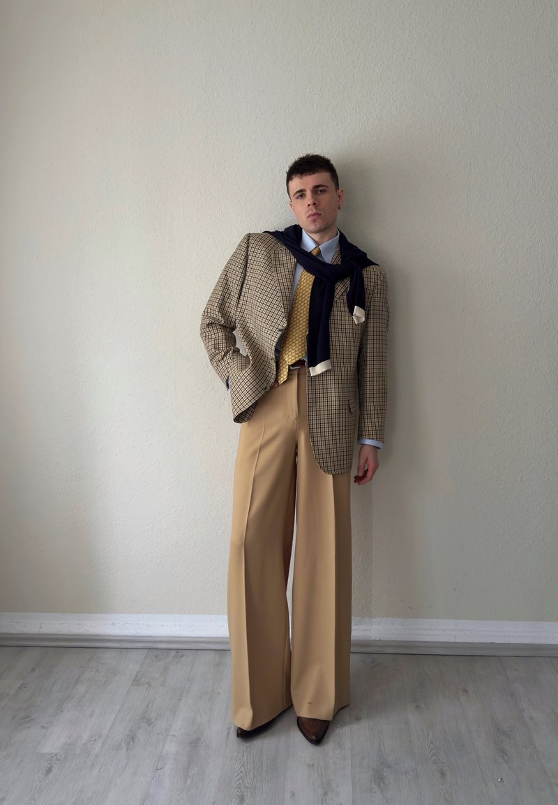 Junger Mann in beigefarbenen, weit geschnittenen Hosen, gelber Weste, kariertem Blazer und einem dunkelblauen Pullover, der über die Schultern gelegt ist, steht vor einer schlichten Wand.