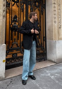 Black blazer over a black top, light blue wide-leg jeans, and black leather shoes standing near an ornate wrought iron gate.