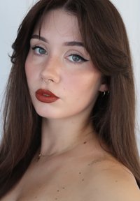 Porträtt av en person med långt, mörkbrunt hår, klara blå ögon och fräknar. Har en gyllene septumring och brun matt läppstift.