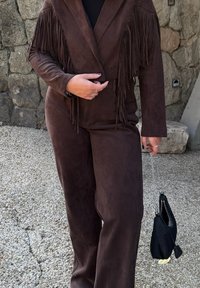 Brązowy zamszowy strój z marynarką z frędzlami i spodniami o szerokich nogawkach, w połączeniu z małą czarną torebką. Teksturowane wykończenie podkreśla atrakcyjność wizualną.