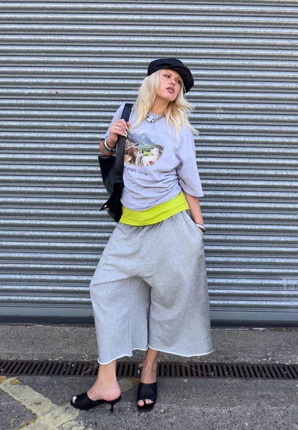 Person trägt graue Hose mit weitem Bein, Grafik-T-Shirt, schwarze Kappe, schwarze Sandalen mit hohem Absatz und trägt eine schwarze Tasche, steht vor einem Metallrollladen.