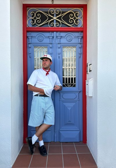 Puerta de madera azul con adornos negros ornamentales. Persona viste una camisa blanca, un pañuelo rojo, pantalones cortos a rayas azul claro, zapatos negros y calcetines blancos.