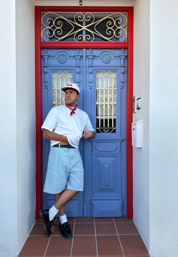 Porta de madeira azul com acentos negros ornamentados. A pessoa veste uma camisa branca, bandana vermelha, calções listrados azul claro, sapatos pretos e meias brancas.
