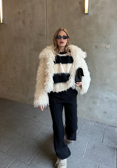 Chaqueta de piel sintética blanca con rayas negras, textura suave, combinada con un suéter de punto a rayas, pantalones negros y zapatos topo.