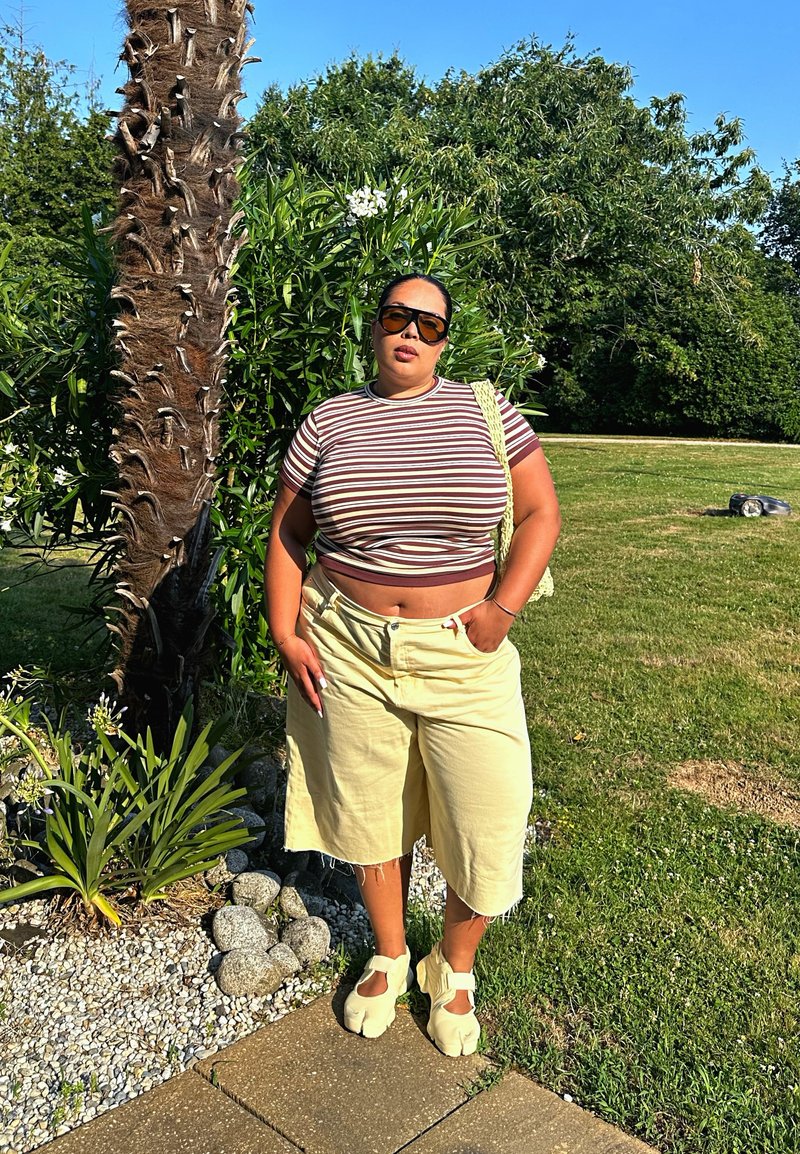 Γυναίκα που φοράει ριγωτό crop top, μπεζ σορτς και παπούτσια με διαχωρισμένα δάχτυλα, στέκεται δίπλα σε μια φοινικιά σε έναν ηλιόλουστο κήπο με πράσινα φυτά και γρασίδι.