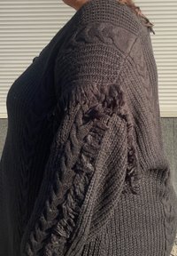 Přerostlý černý pletený svetr se vzory copánkového pletení, texturovanou látkou a třásněmi na okrajích rukávů. Boční pohled.