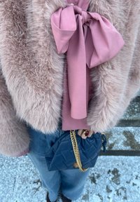 Femme portant un manteau en fausse fourrure rose pâle et un chemisier avec un grand nœud, jean bleu, tenant un sac matelassé bleu marine avec une bandoulière chaîne dorée.