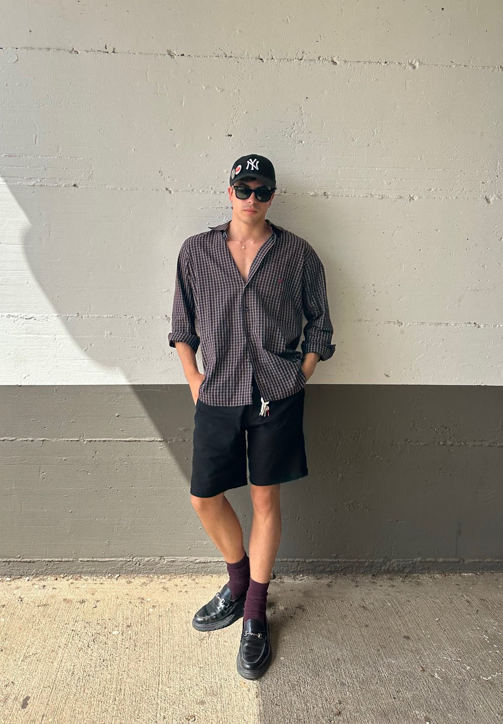 Uomo che indossa una camicia a quadri con bottoni, pantaloni corti neri, mocassini neri e calzini bordeaux, in piedi contro un muro grigio texturizzato.