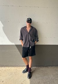 Uomo che indossa una camicia a quadri con bottoni, pantaloni corti neri, mocassini neri e calzini bordeaux, in piedi contro un muro grigio texturizzato.