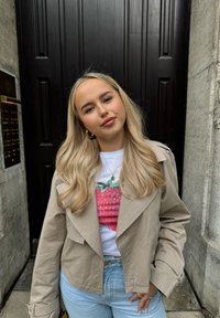 Młoda kobieta z długimi blond włosami, w beżowej kurtce, białym t-shircie w truskawki i jasnoniebieskich dżinsach, stojąca przed ciemnymi drewnianymi drzwiami.