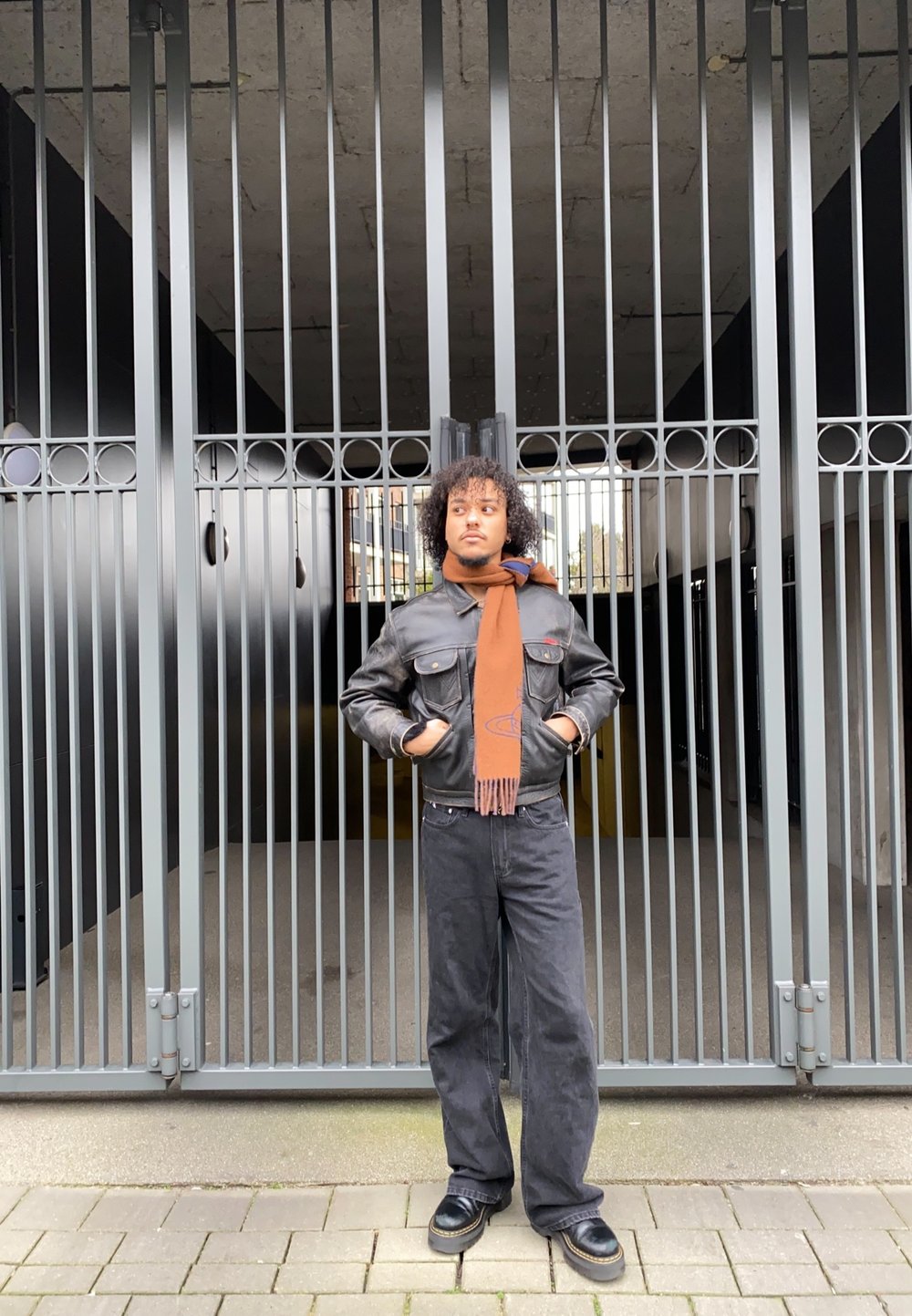 Black leather jacket with two front pockets, brown scarf, black wide-leg jeans, and chunky black shoes in front of a metal gate.