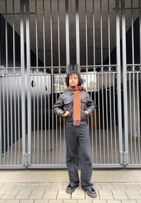 Black leather jacket with two front pockets, brown scarf, black wide-leg jeans, and chunky black shoes in front of a metal gate.