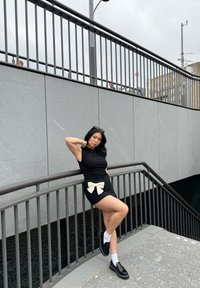 Robe noire cintrée avec un nœud blanc à la taille, associée à des chaussures noires et des chaussettes blanches. L'arri�ère-plan présente des murs et des rampes gris.