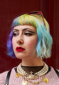 Γυναίκα με κοντά μαλλιά σε χρώματα του ουράνιου τόξου, έντονο eyeliner με φτερούγες, φωτεινά κόκκινα χείλη, ρινικά σκουλαρίκια, πέρλες γύρω από τον λαιμό και ροζ πουλόβερ σε σκούρο κόκκινο φόντο.