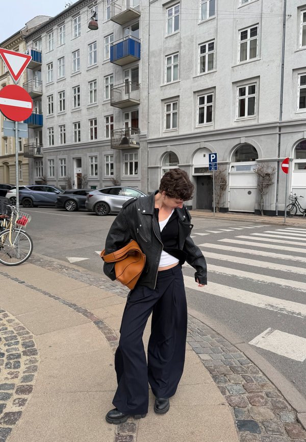 Person iført svart skinnjakke og vide svarte bukser står ved et fotgjengerfelt på en bygate med parkerte biler og sykler i nærheten.