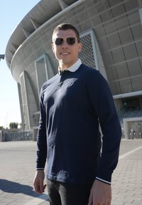 Marinblå langærmet polo-shirt med hvid krave, parret med sorte bukser. Solbriller reflekterer omgivelserne; moderne stadion i baggrunden.