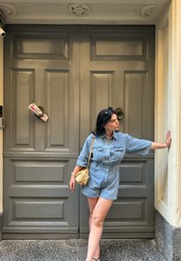 Τζιν ολόσωμο φόρεμα με κοντά μανίκια και δύο τσέπες στο στήθος, σε ανοιχτό μπλε χρώμα. Υφαντή τσάντα και μαύρες σαγιονάρες. Γκρίζες πάνελ διπλές πόρτες στο παρασκήνιο.