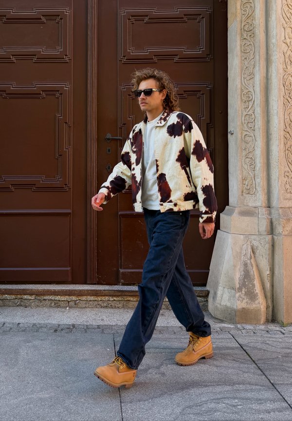 Un hombre con gafas de sol, una chaqueta estampada, vaqueros oscuros y botas de color tostado pasa junto a una puerta marrón ornamentada.
