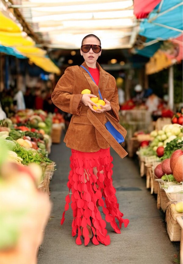 Kvinde iført oversize brun jakke og rød nederdel med cirkulære udskæringer, holder citroner og står i en farverig udendørs markedsgang.