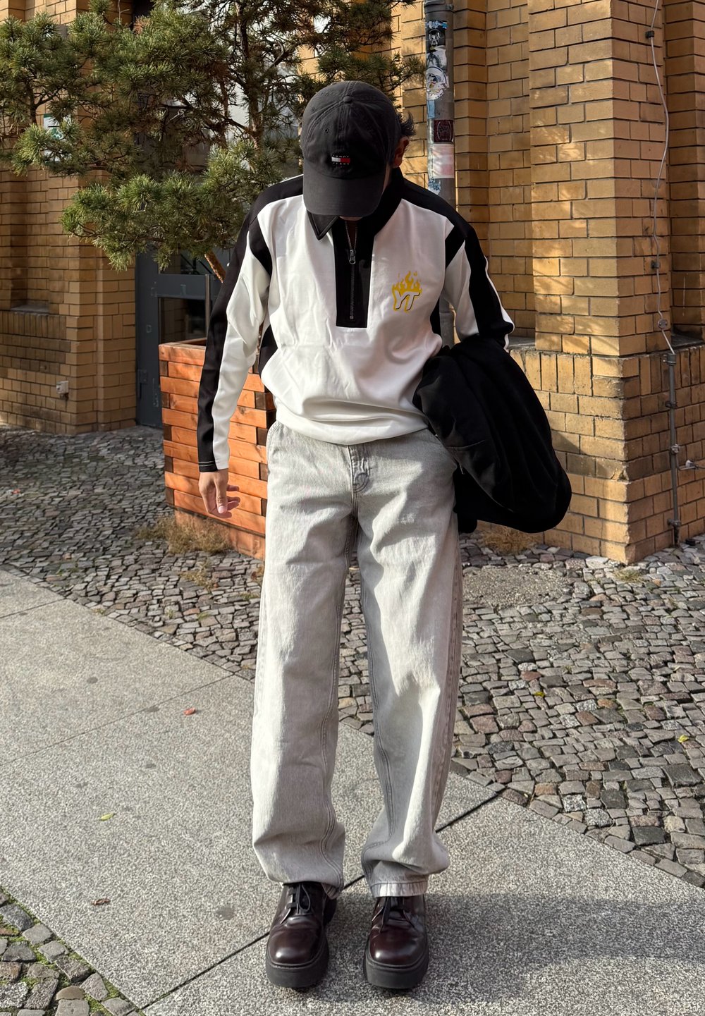 Biała i czarna kurtka na zamek z żółtym logo płomienia, jasnoszare jeansy o szerokich nogawkach i ciemnobrązowe buty sznurowane, trzymająca czarną kurtkę.