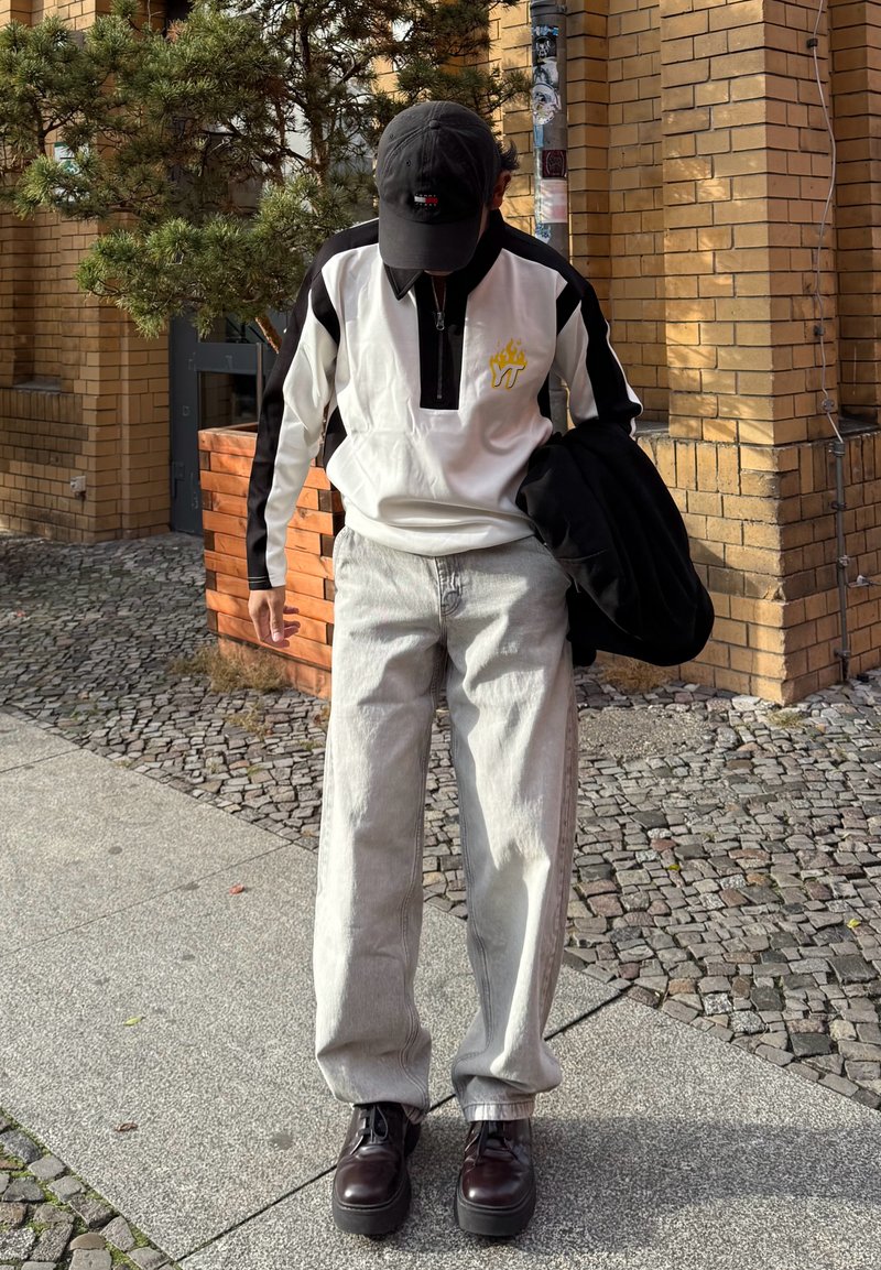 White and black zip-up jacket with a yellow flame logo, light gray wide-leg jeans, and dark brown lace-up shoes, holding a black jacket.