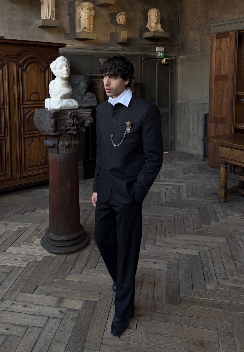 Man in black suit with white collar and decorative pin stands near classical marble bust on wooden pedestal in historic room.