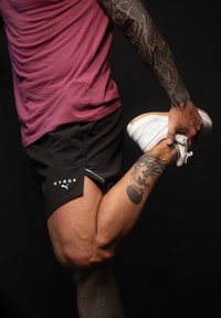 Černé sportovní šortky s logem "HYROX" v kombinaci s bílými sportovními botami a přiléhavým vínovým tričkem. Viditelné tetování na paži.