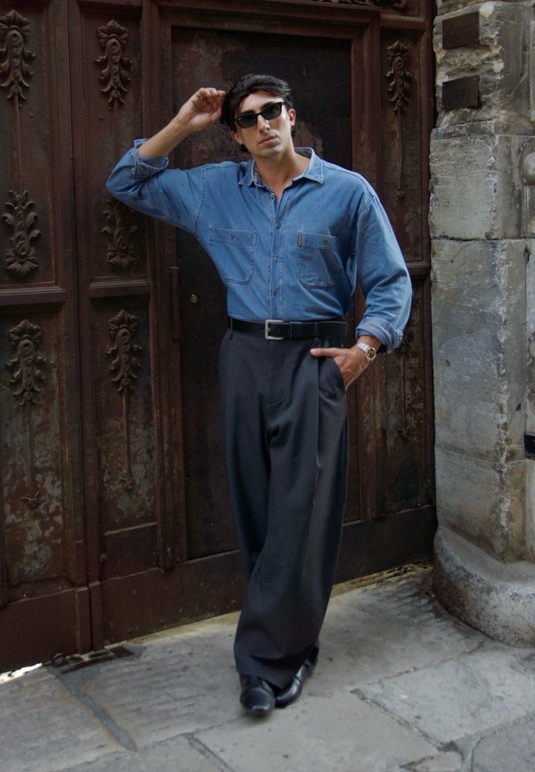 Hombre con gafas de sol, vestido con una camisa vaquera azul y pantalones negros de pierna ancha, apoyado de manera casual contra una puerta de madera ornamentada.