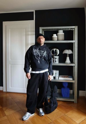 Sudadera negra con gráficos blancos, pantalones cargo negros, zapatillas deportivas plateadas y una bolsa negra. El fondo presenta estanterías con objetos decorativos.