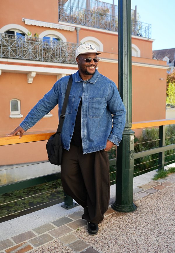 Hombre con gafas de sol, chaqueta vaquera y gorra, sonriendo mientras se apoya en una barandilla junto a un canal en una zona urbana soleada.