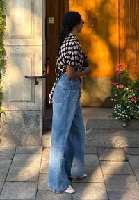Femme aux cheveux tressés portant un chemisier à pois et un jean large bleu, debout près d'une porte en bois à côté de fleurs sur des marches en pierre.