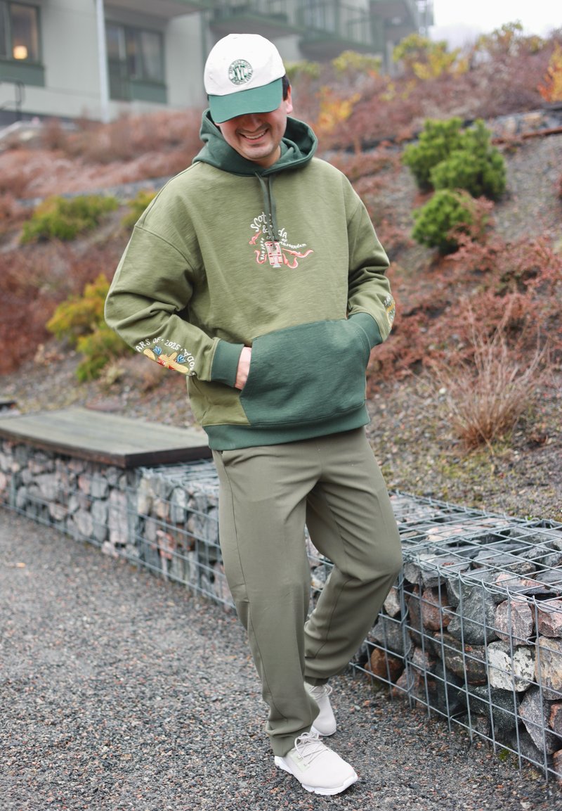 Mann in grünem Hoodie, olivfarbenen Hosen, weißen Sneakers und weiß-grüner Mütze steht auf einem Kiesweg neben einer Steinmauer und Sträuchern, lächelt und hat die Hände in den Taschen.