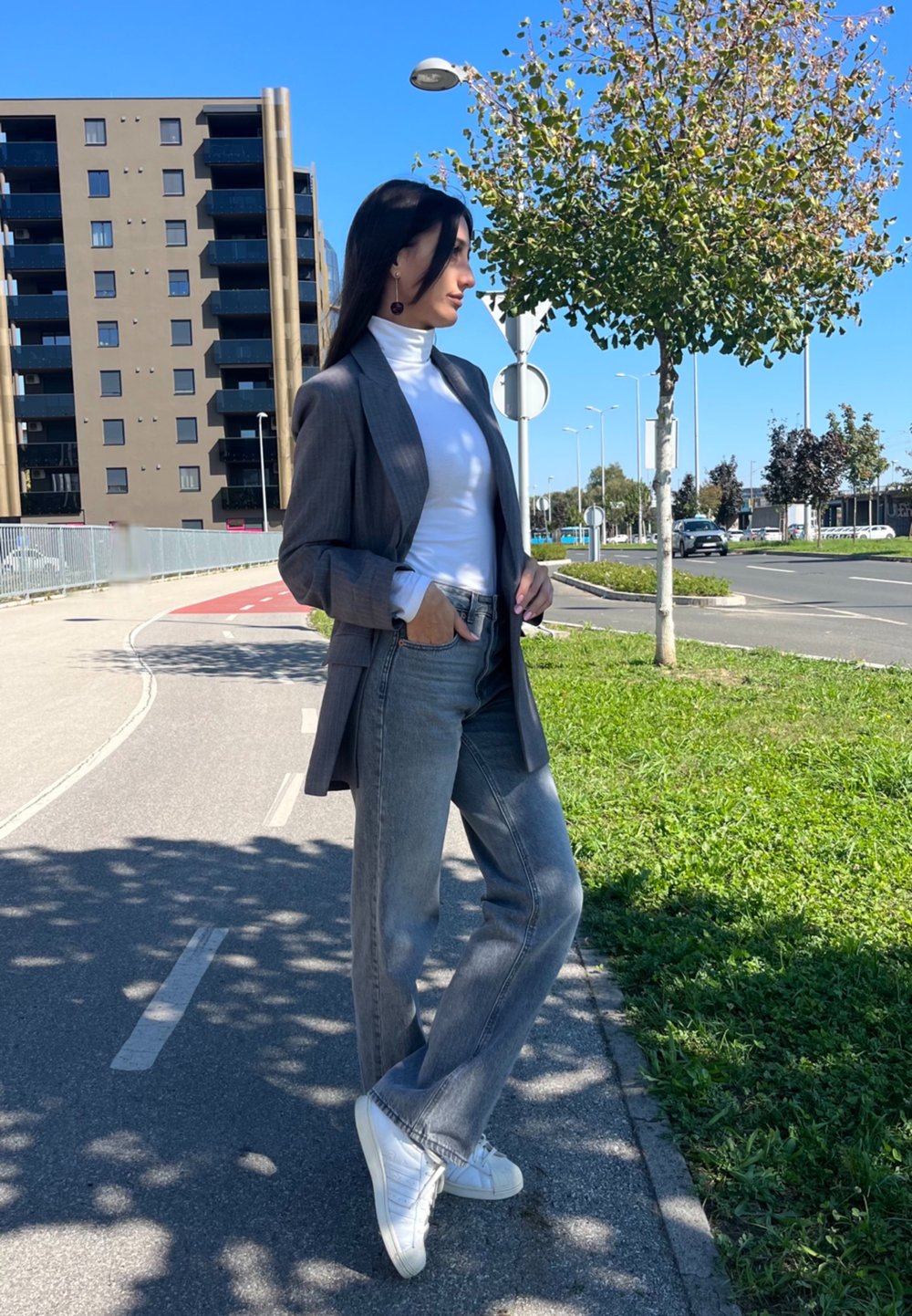 Γκρίζα καρό σακάκι πάνω από λευκή μπλούζα με ζιβάγκο, συνδυασμένο με ανοιχτό γκρι τζιν και λε�υκά αθλητικά παπούτσια. Υπαίθριο σκηνικό με δέντρα και κτίρια.