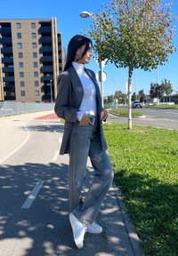 Γκρίζα καρό σακάκι πάνω από λευκή μπλούζα με ζιβάγκο, συνδυασμένο με ανοιχτό γκρι τζιν και λευκά αθλητικά παπούτσια. Υπαίθριο σκηνικό με δέντρα και κτίρια.