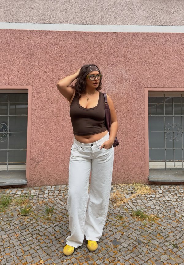 Vrouw met bril en hoofdband, draagt een bruin hemdje, witte broek, gele schoenen, staand tegen een roze gestructureerde muur met twee ramen.