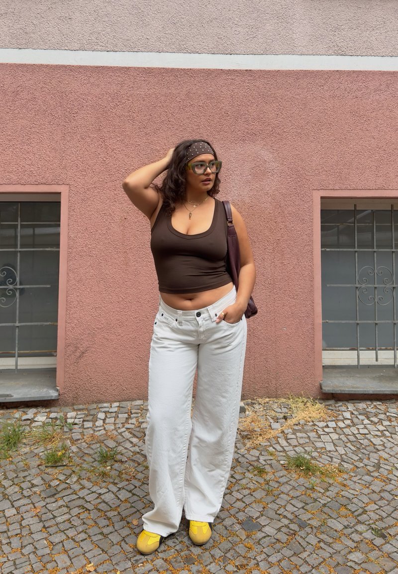 Femme avec des lunettes et un bandeau portant un débardeur marron, un pantalon blanc, des chaussures jaunes, debout contre un mur rose texturé avec deux fenêtres.