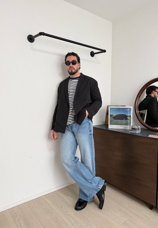 Mann med solbriller, svart blazer, stripete skjorte og lyseblå jeans lener seg mot en hvit vegg ved siden av en kommode i tre med et rundt speil.