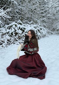 Ung kvinde iført en mønstret sweater og lang bordeaux nederdel knæler i sneen og holder et hvidt tørklæde, med snedækkede træer i baggrunden.