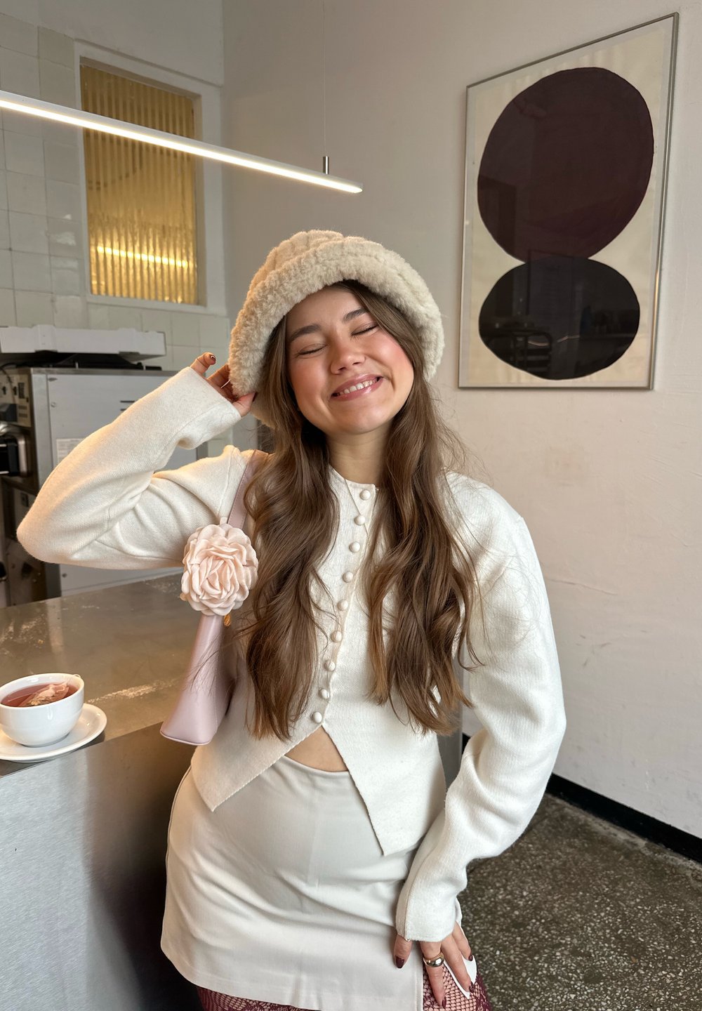 Jeune femme souriante les yeux fermés, portant un haut blanc boutonné, une jupe beige, un chapeau duveteux et un sac rose avec une fleur, debout près d'un comptoir.