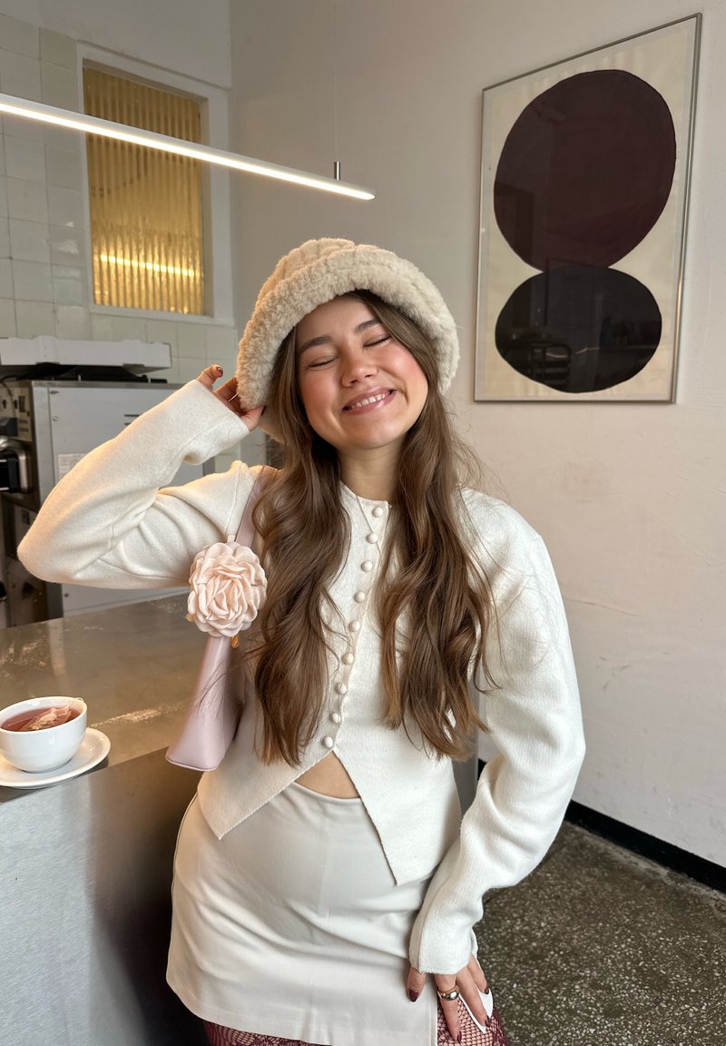 Jeune femme souriante les yeux fermés, portant un haut blanc boutonné, une jupe beige, un chapeau duveteux et un sac rose avec une fleur, debout près d'un comptoir.