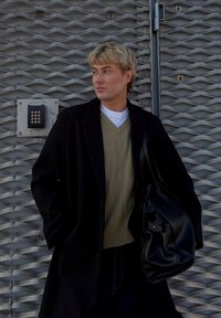 Cappotto nero sopra un maglione verde, jeans in denim e una borsa nera in pelle. Lo sfondo presenta una superficie metallica strutturata con una tastiera.