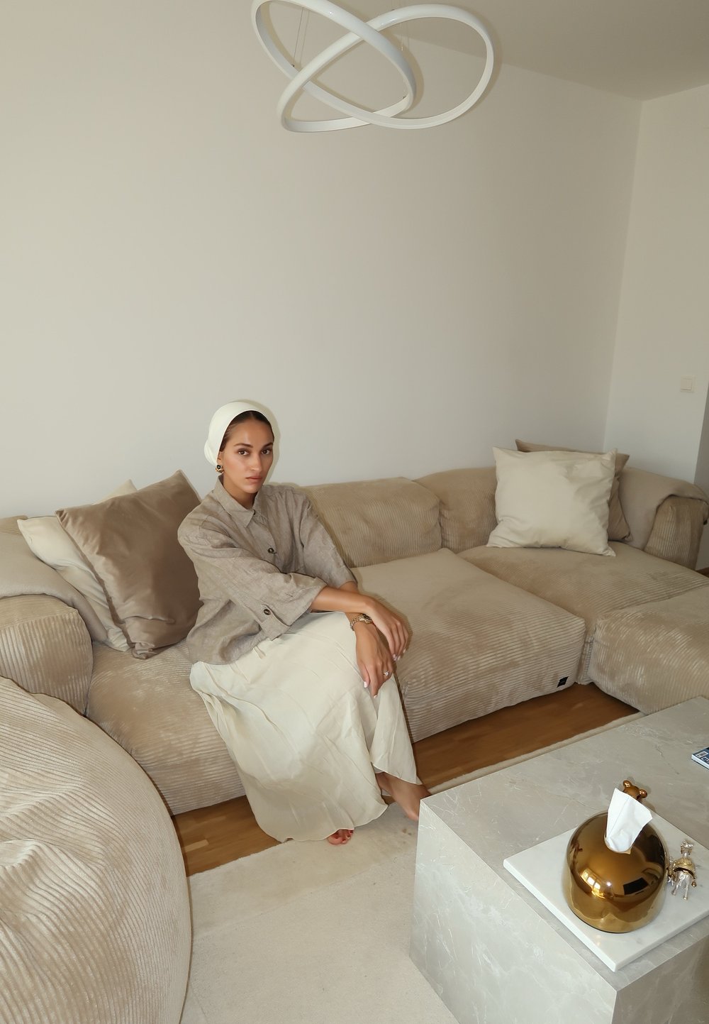 Bežani kordurov kauč s odgovarajućim jastucima, žena u svijetloj lanenoj haljini i zlatni držač maramica na bijelom mramornom stolu. Minimalistički dekor.