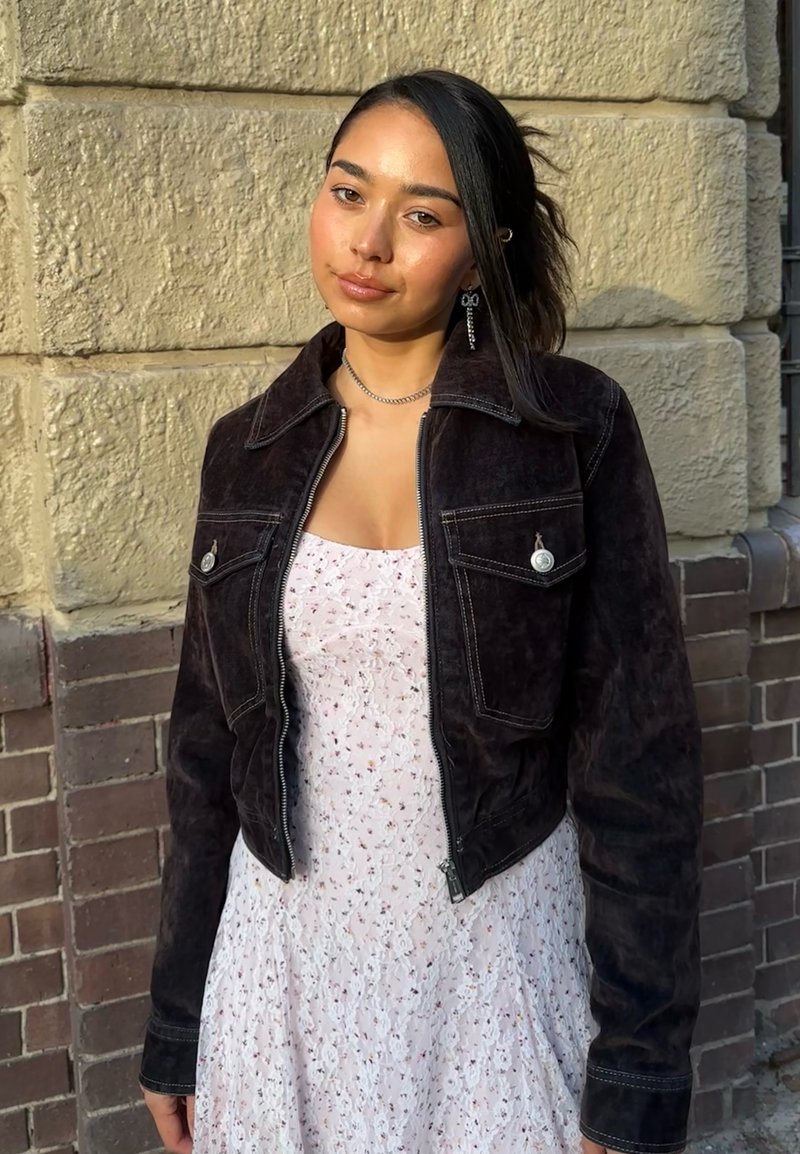 Jeune femme aux cheveux foncés porte une veste noire sur une robe fleurie claire, debout contre un mur de pierre texturé sous une lumière douce.