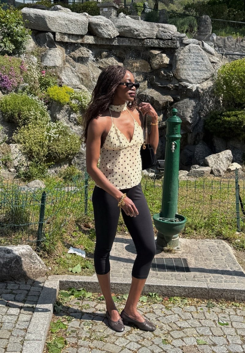 Γυναίκα που φοράει τοπάκι με πουά και μαύρο capri κολάν στέκεται κοντά σε ένα πράσινο σιντριβάνι με βραχώδη κήπο στο φόντο.