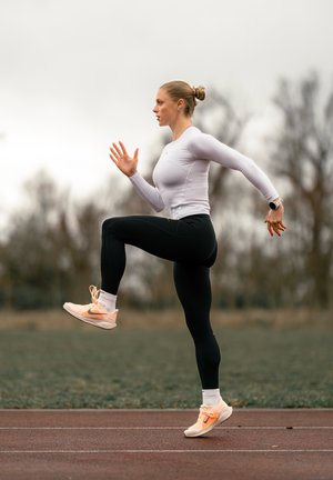 Conjunto deportivo con camiseta blanca ceñida de manga larga y leggings negros. Calzado: zapatillas rosa claro con detalles en naranja. Corriendo en una pista.