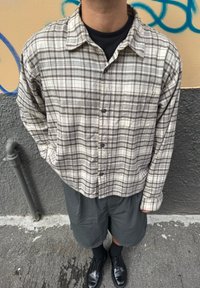 Beige és szürke kockás ing, laza szabásban, sötét rövidnadrággal és fekete cipővel párosítva, egy texturált szürke háttér előtt áll.