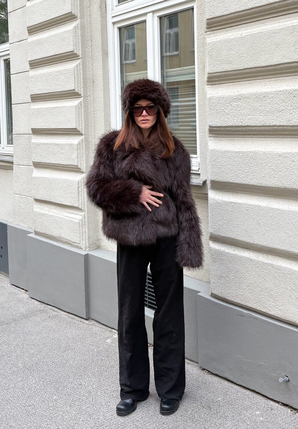 Jachetă maro din blană artificială cu o pălărie asortată, croială largă, purtată cu pantaloni negri cu picioare largi și ochelari de soare rotunzi, pe fundalul unui zid texturat.